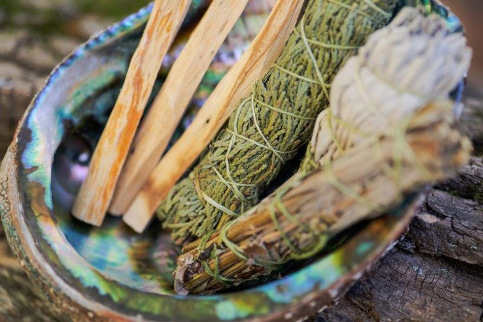 Bundles of sage and other herbs beside palo santo sticks sitting inside a shell