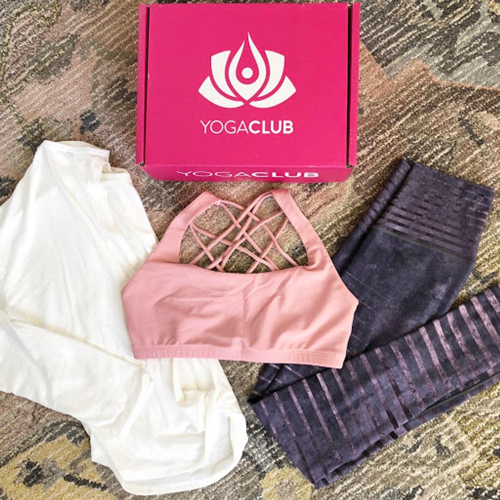 A pink sports bra, purple yoga pants, and a white tank are spread out next to a YogaClub box