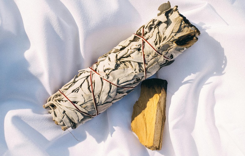 Bundles of sage and other herbs beside palo santo sticks sitting inside a shell